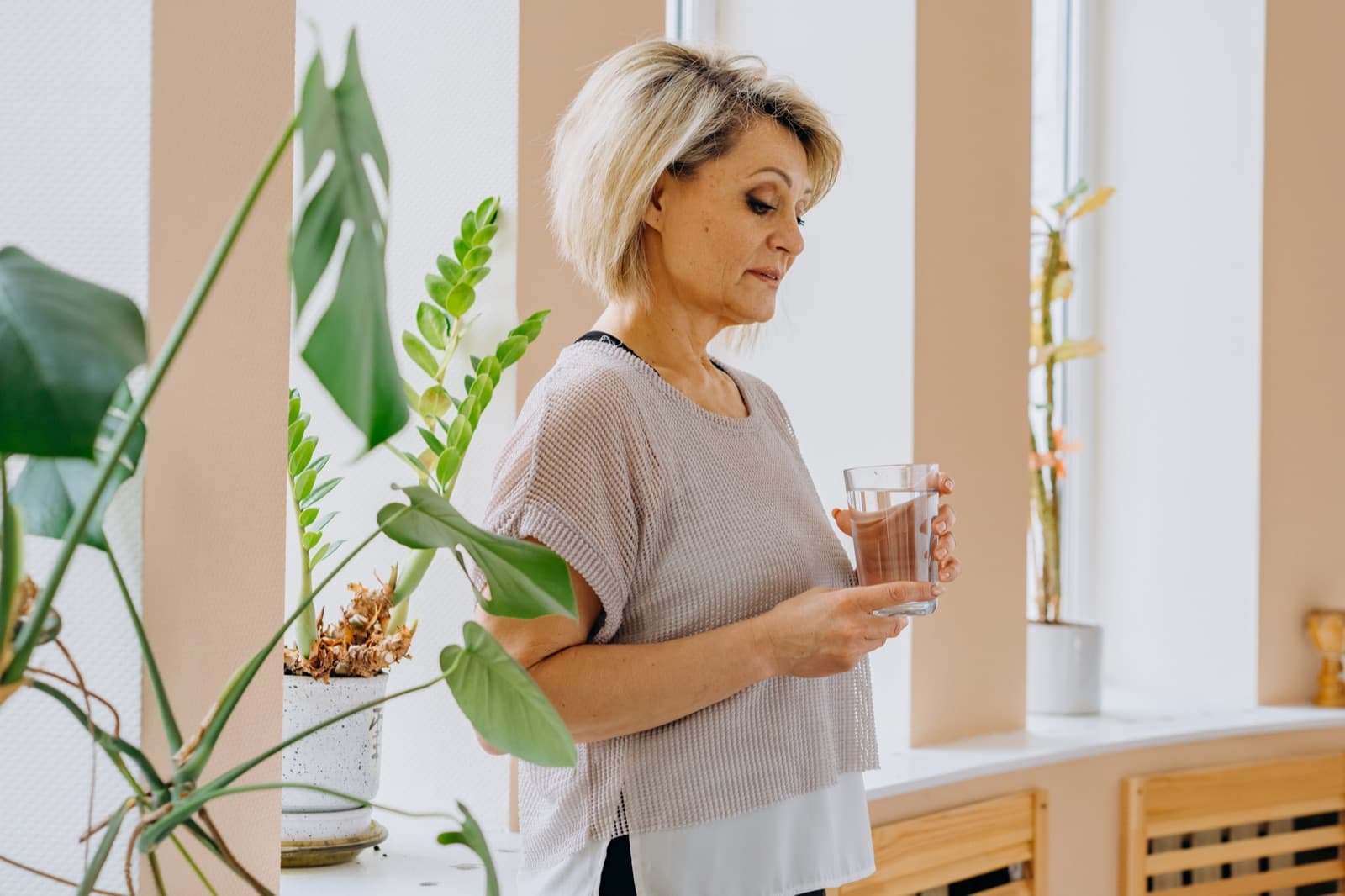 Woman in her 50s standing quietly by a window holding a glass of water, looking reflective