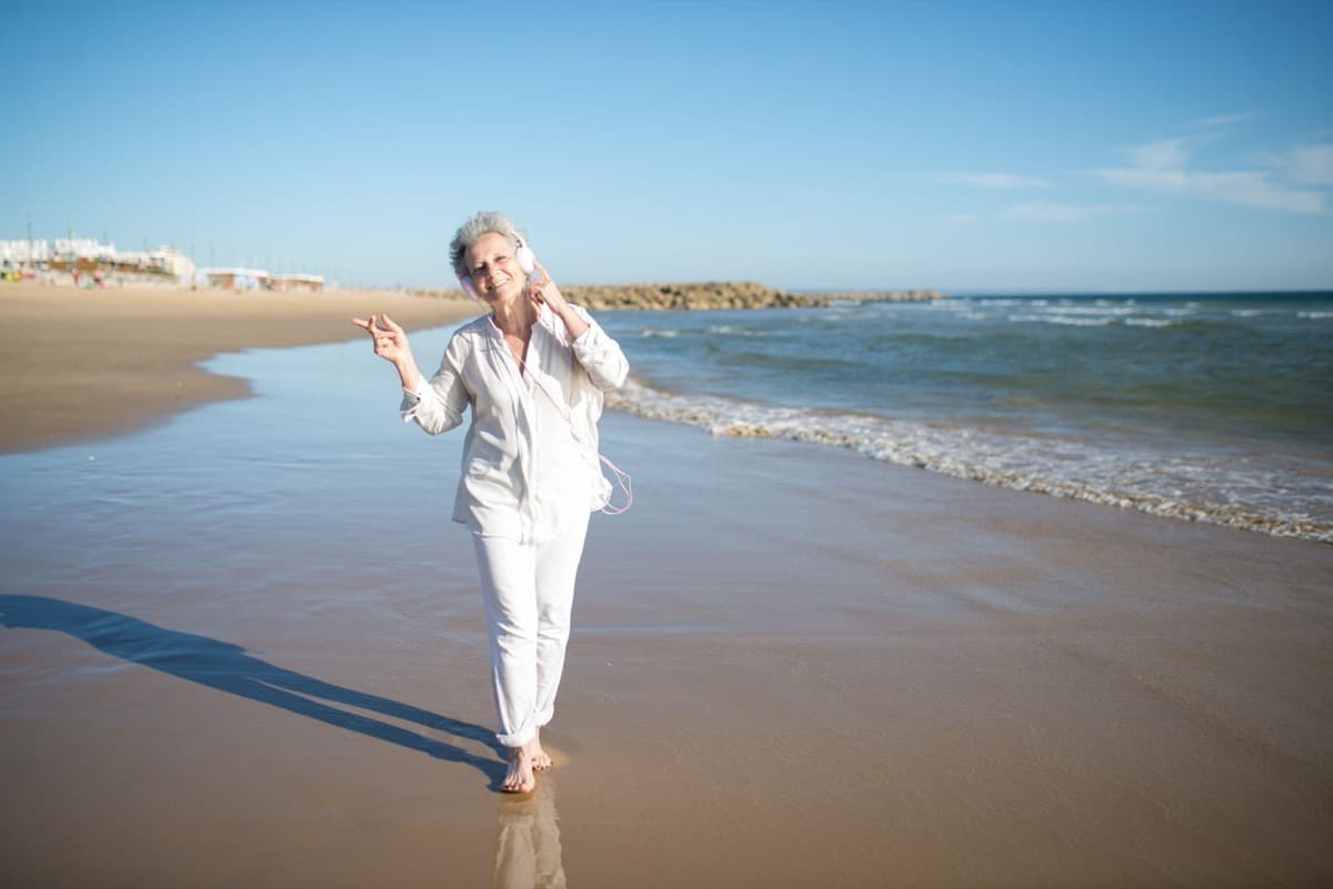 Older woman walking along a sunny beach, smiling and listening to music